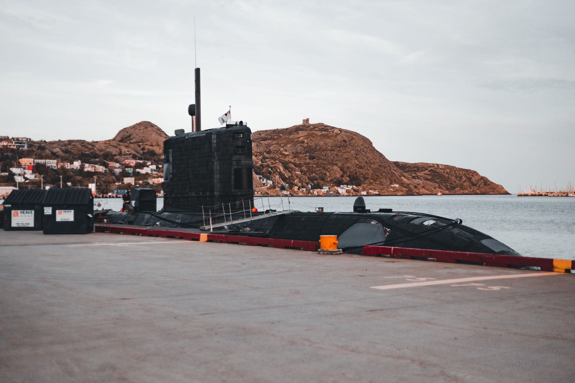 A submarine docked at an urban waterfront with scenic hills in the background.