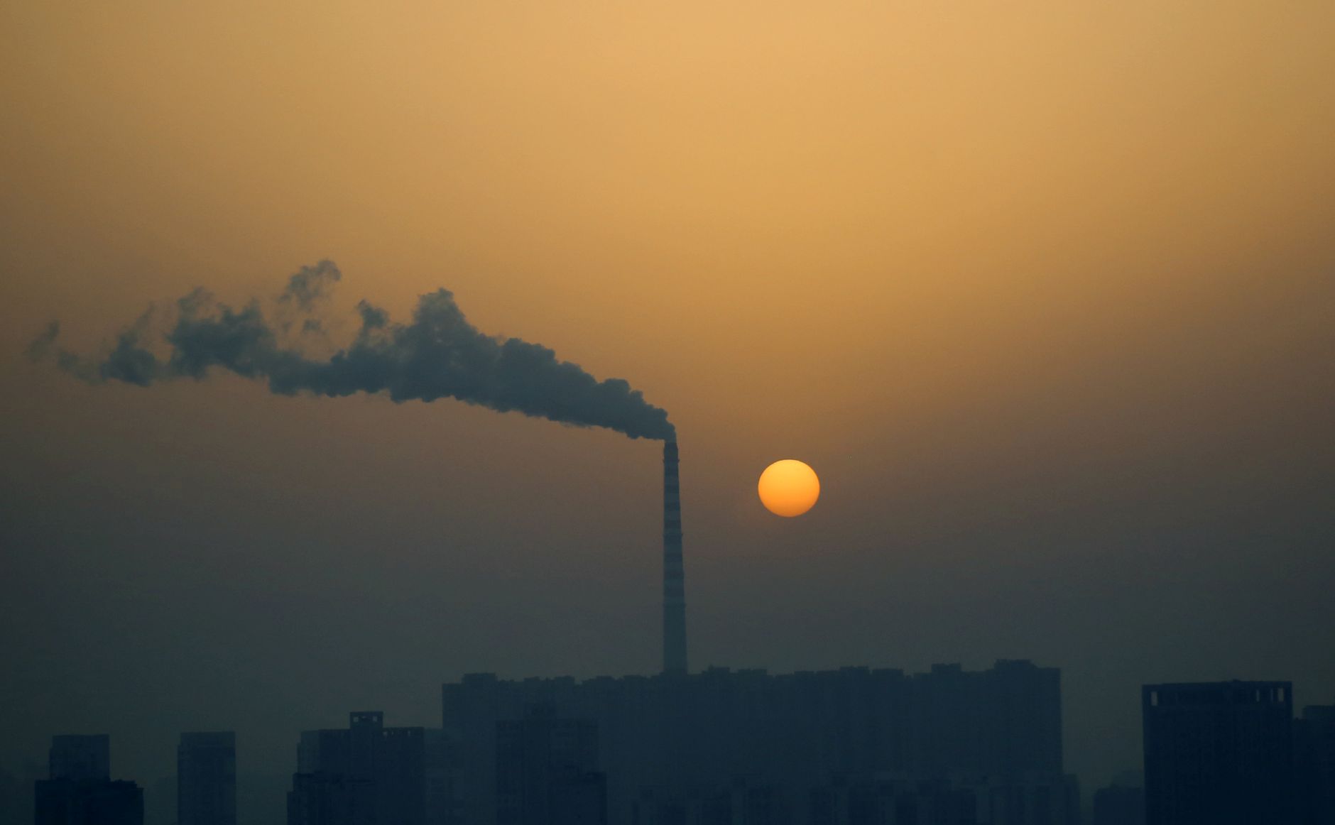 Silhouette of cityscape and smokestack emitting smoke against a reddish sunrise sky.