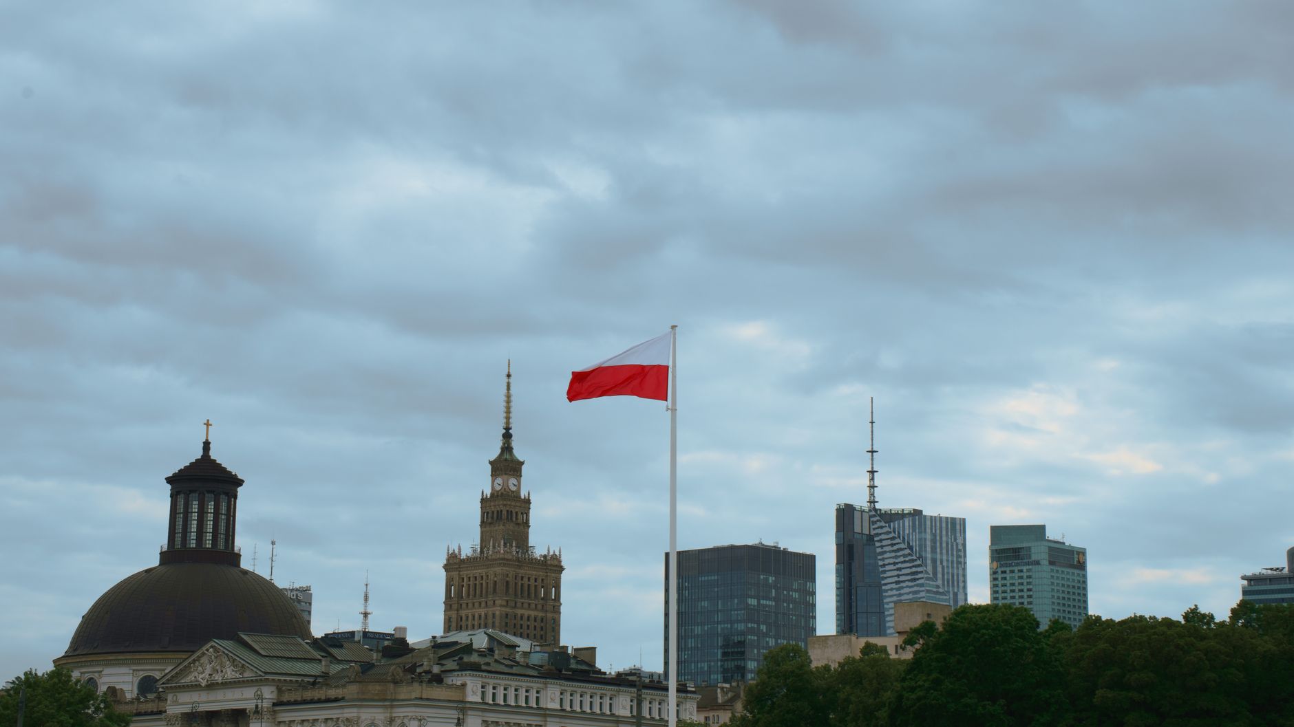 Skyline of Warsaw featuring the Polish flag and iconic buildings under a cloudy sky.