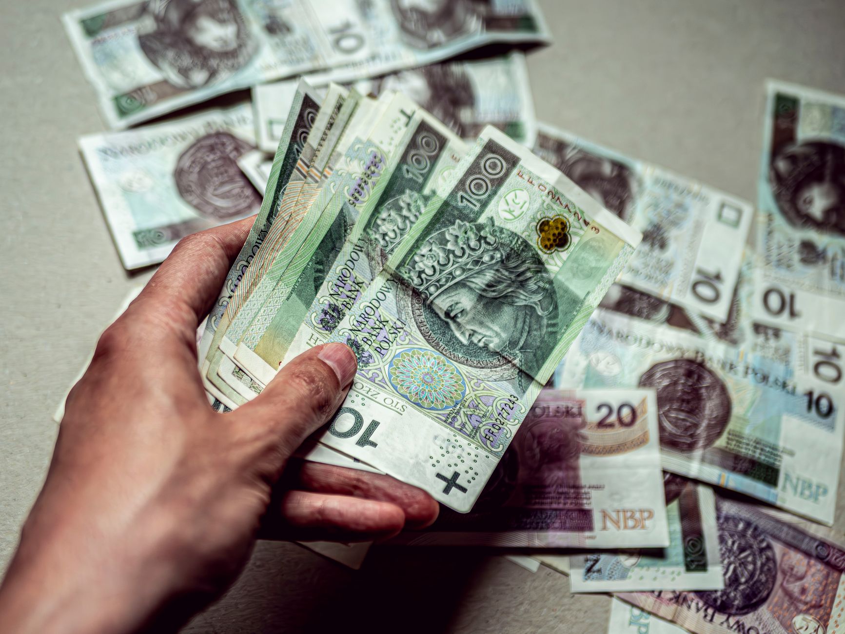 A closeup of a hand holding Polish Zloty banknotes with scattered currency in the background.